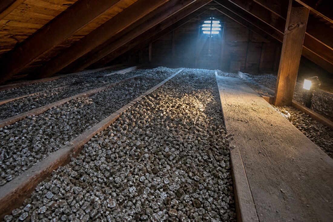 Loose-fill vermiculite insulation filling the spaces between wooden ceiling joists in the attic of an older Fraser Valley home, lit by a work light and daylight from a gable vent